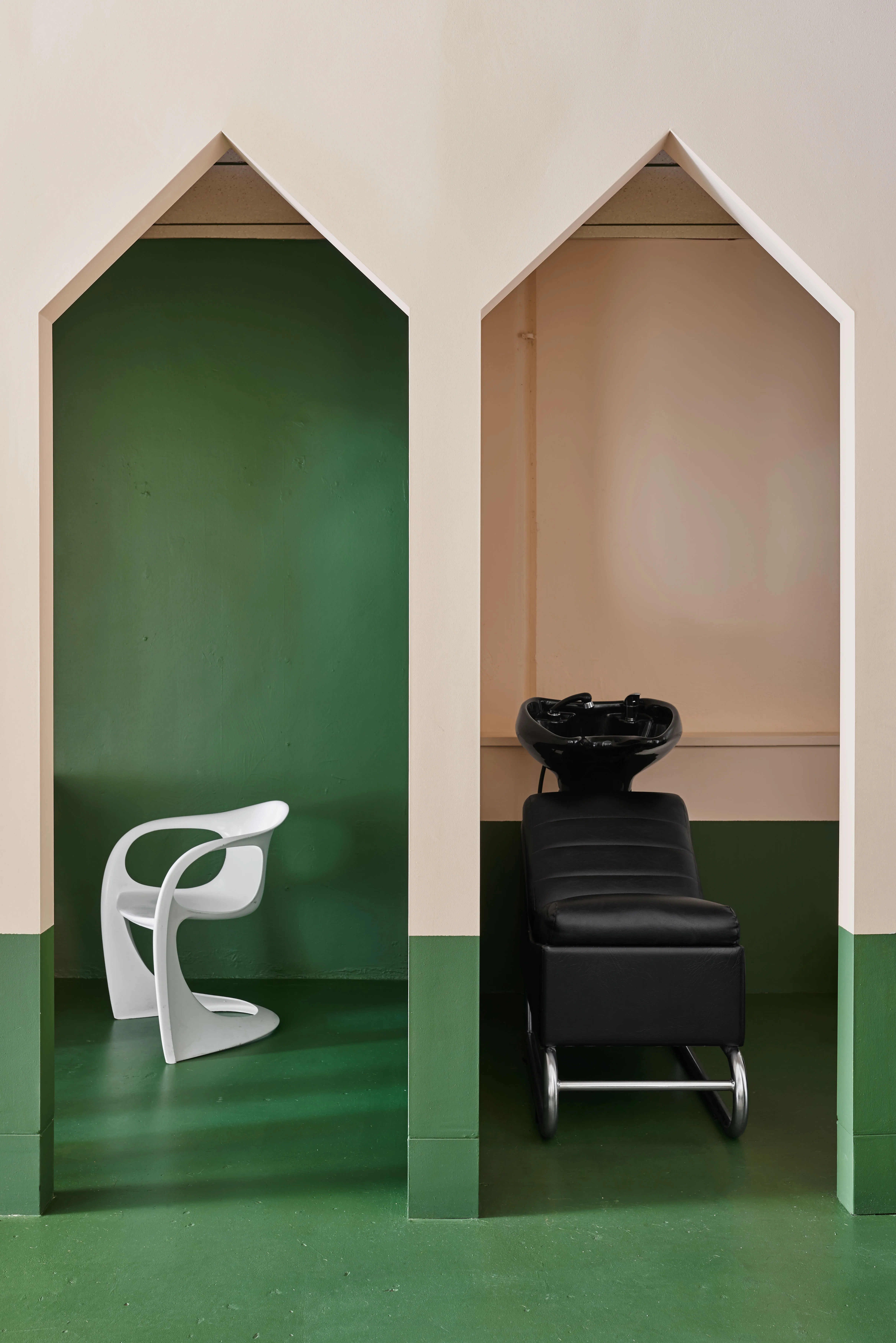 The twin Gothic arch alcoves inside Hues Hair — a white sculptural cantilever chair in the green-painted alcove, a black leather shampoo chair in the peach-toned alcove